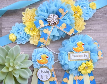 May include: A baby shower sash and two pins featuring light blue and yellow fabric flowers. The sash has a central rhinestone accent and a blue ribbon with yellow rubber duck prints. The pins feature rubber ducks and text reading Mommy to Be and Daddy to Be.