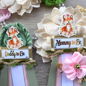 May include: Baby shower rosette pins with a fox theme. The pins feature a fabric flower base, a ribbon, and a small sign that says "Daddy to Be" and "Mommy to Be". The pins are decorated with pearls and a fox illustration.