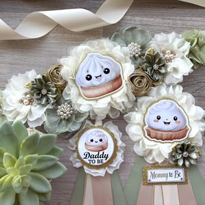 May include: A baby shower sash and pins with a dumpling theme. The sash is white with floral accents in white, green, and gold. The pins say "Daddy to Be" and "Mommy to Be" and feature a cute dumpling graphic.