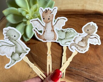 May include: Four cupcake toppers featuring cartoon dragons. The dragons are brown, green, and gray with white wings. The toppers are attached to wooden sticks.