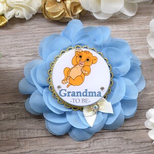 Pu&ograve; includere: Spilla a forma di fiore azzurro chiaro con un centro bordato d'oro raffigurante un cucciolo di leone dei cartoni animati e la scritta "Nonna -TO BE-". La spilla ha un fiocco bianco con un dettaglio di strass.