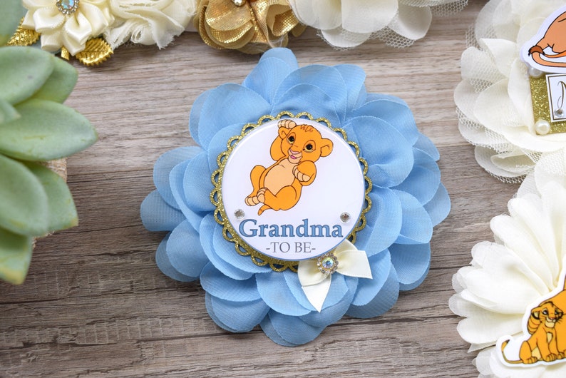Pu&ograve; includere: Spilla a forma di fiore azzurro chiaro con un centro bordato d'oro raffigurante un cucciolo di leone dei cartoni animati e la scritta "Nonna -TO BE-". La spilla ha un fiocco bianco con un dettaglio di strass.