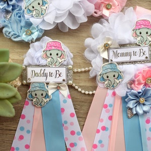 May include: Two baby shower sashes with blue and pink ribbons, white polka dots, and a silver glitter background. The sashes have a white flower with a blue or pink elephant wearing a hat and the text "Daddy to Be" or "Mommy to Be".