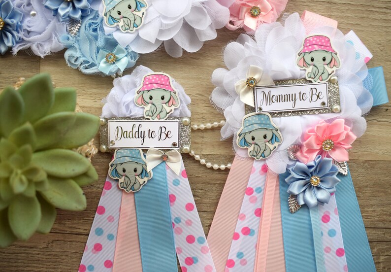 May include: Two baby shower sashes with blue and pink ribbons, white polka dots, and a silver glitter background. The sashes have a white flower with a blue or pink elephant wearing a hat and the text "Daddy to Be" or "Mommy to Be".