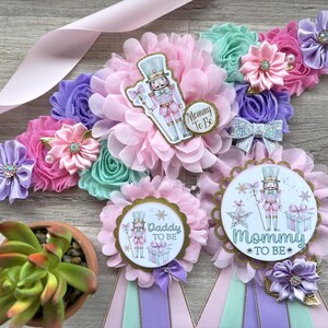 May include: A set of baby shower decorations featuring a pink satin sash adorned with fabric flowers in shades of pink, purple, and mint green. The decorations include rosettes with a nutcracker design and the text "Mommy To Be" and "Daddy To Be".