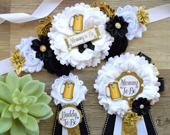 May include: A set of three white and black sashes with gold accents and beer-themed embellishments. The sashes have the text "Mommy to Be" and "Daddy to Be" printed on them.