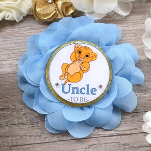 Pu&ograve; includere: Un fiore di tessuto azzurro con un centro circolare raffigurante un cucciolo di leone dei cartoni animati e la scritta "Uncle -TO BE-". Il fiore ha pi&ugrave; strati di petali e un bordo glitterato dorato.