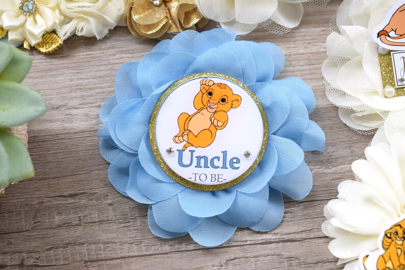 Pu&ograve; includere: Un fiore di tessuto azzurro con un centro circolare raffigurante un cucciolo di leone dei cartoni animati e la scritta "Uncle -TO BE-". Il fiore ha pi&ugrave; strati di petali e un bordo glitterato dorato.