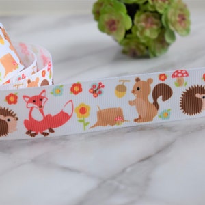 May include: A white grosgrain ribbon featuring a repeating pattern of orange foxes, brown hedgehogs, and light brown squirrels. The design includes small yellow flowers, red mushrooms, and acorns scattered across the fabric surface in a continuous print.