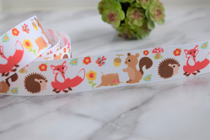 May include: A white grosgrain ribbon featuring a repeating pattern of orange foxes, brown hedgehogs, and light brown squirrels. The design includes small yellow flowers, red mushrooms, and acorns scattered across the fabric surface in a continuous print.
