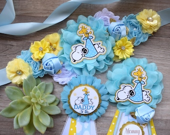 May include: A baby shower sash and two pins featuring Snoopy and Woodstock. The sash has light blue and yellow fabric flowers with a central Snoopy graphic. One pin says Daddy-To-Be and the other features Snoopy and Woodstock.