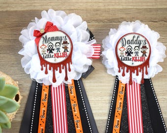 May include: Two white flower-shaped sashes with black, orange, and red ribbons. The sashes have a red circle with white text that reads "Mommy's little killer" and "Daddy's little killer". The circles have images of horror movie characters.