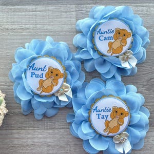Pu&ograve; includere: Tre accessori per capelli a forma di fiore in tessuto azzurro. Ogni fiore ha un centro circolare con un cucciolo di leone dei cartoni animati e le parole "Zia" seguite da un nome. Piccoli fiocchi bianchi e finiture dorate.