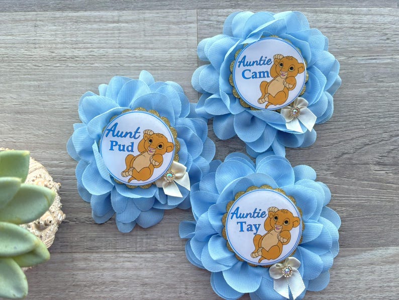 Pu&ograve; includere: Tre accessori per capelli a forma di fiore in tessuto azzurro. Ogni fiore ha un centro circolare con un cucciolo di leone dei cartoni animati e le parole "Zia" seguite da un nome. Piccoli fiocchi bianchi e finiture dorate.