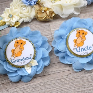 Pu&ograve; includere: Spille a forma di fiore in tessuto azzurro con l'immagine di un cucciolo di leone dei cartoni animati. Una spilla dice "Nonna -TO BE-" e l'altra dice "Zio -TO BE-". Le spille hanno finiture dorate e sono decorate con piccoli strass e un fiocco.