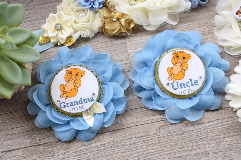 Pu&ograve; includere: Spille a forma di fiore in tessuto azzurro con l'immagine di un cucciolo di leone dei cartoni animati. Una spilla dice "Nonna -TO BE-" e l'altra dice "Zio -TO BE-". Le spille hanno finiture dorate e sono decorate con piccoli strass e un fiocco.