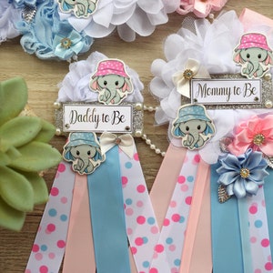 May include: Two baby shower sashes with blue and pink ribbons, white polka dots, and elephant designs. One sash says "Mommy to Be" and the other says "Daddy to Be".