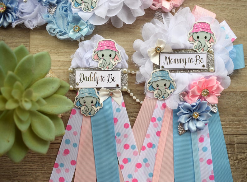 May include: Two baby shower sashes with blue and pink ribbons, white polka dots, and elephant designs. One sash says "Mommy to Be" and the other says "Daddy to Be".