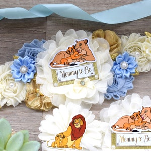 Pu&ograve; includere: Un nastro di raso azzurro con una fascia floreale con fiori di tessuto avorio, oro e blu. La fascia ha un cartello "Mommy to Be" con un'illustrazione di una leonessa e del suo cucciolo.