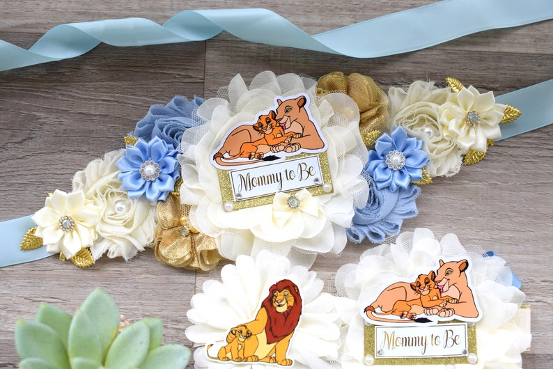 Pu&ograve; includere: Un nastro di raso azzurro con una fascia floreale con fiori di tessuto avorio, oro e blu. La fascia ha un cartello "Mommy to Be" con un'illustrazione di una leonessa e del suo cucciolo.