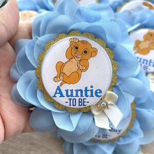 Pu&ograve; includere: Bomboniere per baby shower a forma di fiori azzurri. Ogni bomboniera presenta una grafica di Simba con il testo "Auntie -TO BE-" su sfondo bianco, circondata da un bordo glitterato dorato. &Egrave; attaccato un piccolo fiocco bianco con un cristallo.
