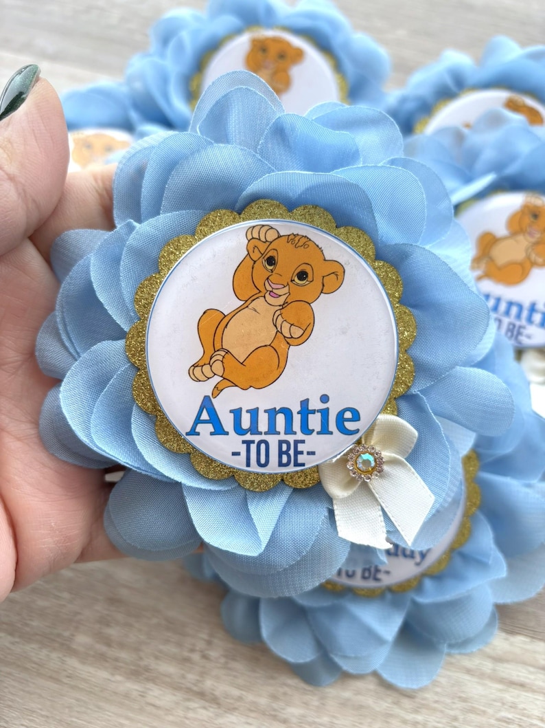 Pu&ograve; includere: Bomboniere per baby shower a forma di fiori azzurri. Ogni bomboniera presenta una grafica di Simba con il testo "Auntie -TO BE-" su sfondo bianco, circondata da un bordo glitterato dorato. &Egrave; attaccato un piccolo fiocco bianco con un cristallo.