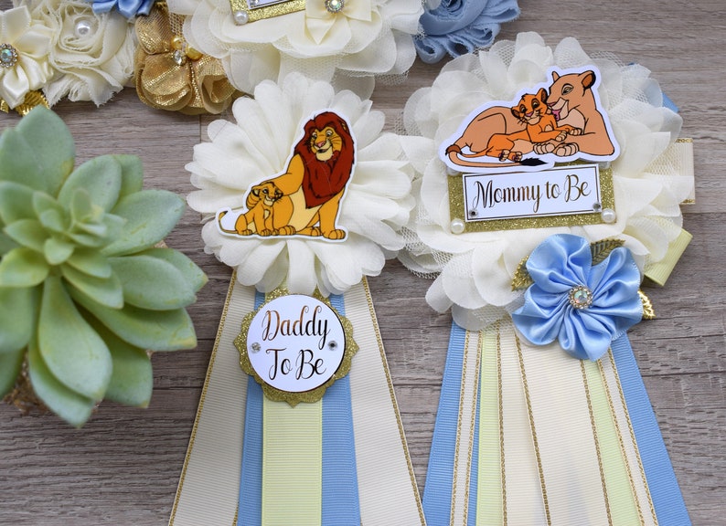Pu&ograve; includere: Decorazioni per baby shower a tema Il Re Leone. Accenti floreali color crema con nastri blu, gialli e oro. Include badge con la scritta "Daddy To Be" e "Mommy To Be".