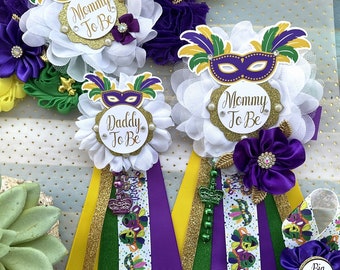 May include: Two white flower corsages with purple and green Mardi Gras themed ribbons. One corsage says "Mommy To Be" and the other says "Daddy To Be".