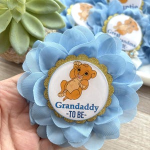 Pu&ograve; includere: Fiore di tessuto azzurro con un centro dorato raffigurante un cucciolo di leone dei cartoni animati e la scritta "Grandaddy -TO BE-". Il fiore ha pi&ugrave; strati di petali ed &egrave; probabilmente un oggetto decorativo per un baby shower.