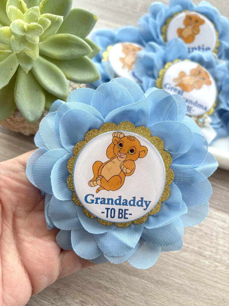 Pu&ograve; includere: Fiore di tessuto azzurro con un centro dorato raffigurante un cucciolo di leone dei cartoni animati e la scritta "Grandaddy -TO BE-". Il fiore ha pi&ugrave; strati di petali ed &egrave; probabilmente un oggetto decorativo per un baby shower.