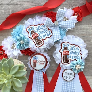 May include: A set of baby shower decorations featuring red, white, and blue accents. The set includes a sash, rosettes, and badges with the text "She's ready to pop!" and "Daddy To Be" and "Mommy To Be" with a Coca-Cola bottle graphic.