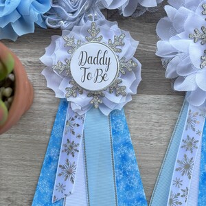 以下が含まれることがあります： 雪の結晶のアクセントが付いた、白とシルバーの「Daddy To Be」ロゼット。ロゼットは白い生地で囲まれ、雪の結晶模様の青と白のリボンが付いています。追加の青とシルバーの花の装飾が見えます。