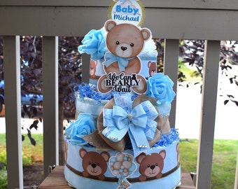 May include: A three-tiered diaper cake decorated with blue and brown teddy bear designs. The top tier features a teddy bear with a blue bow and a sign that says "Baby Michael". The middle tier has a sign that says "We Can Bearly Wait". The bottom tier has a teddy bear holding balloons.