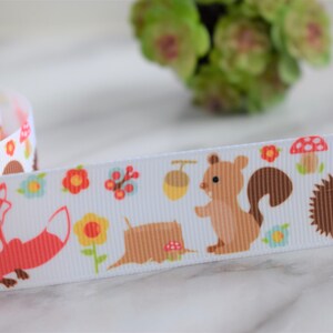 May include: A white grosgrain ribbon featuring a repeating pattern of woodland creatures including orange foxes, brown squirrels, and hedgehogs, interspersed with colourful flowers, small red mushrooms, and brown acorns.