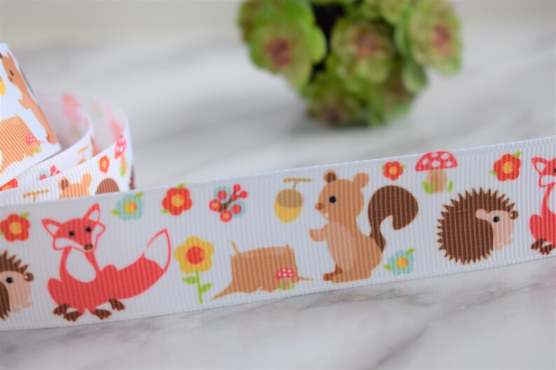 May include: A white grosgrain ribbon featuring a repeating pattern of woodland creatures including orange foxes, brown squirrels, and hedgehogs, interspersed with colourful flowers, small red mushrooms, and brown acorns.