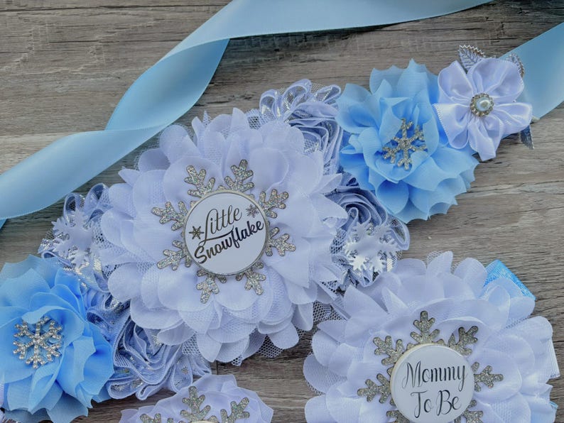 以下が含まれることがあります： 「Little Snowflake」と「Mommy To Be」の文字が入った、水色のリボンと花のサッシュ。白い布と水色の布の花に、銀色の雪の結晶のアクセントが施されており、ベビーシャワーに最適です。