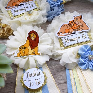 Pu&ograve; includere: Decorazioni per baby shower a tema Il Re Leone. Le decorazioni includono accenti floreali bianchi e dorati, nastri e le frasi "Mommy to Be" e "Daddy to Be". La tavolozza dei colori comprende crema, oro e blu.