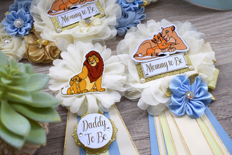 Pu&ograve; includere: Decorazioni per baby shower a tema Il Re Leone. Le decorazioni includono accenti floreali bianchi e dorati, nastri e le frasi "Mommy to Be" e "Daddy to Be". La tavolozza dei colori comprende crema, oro e blu.