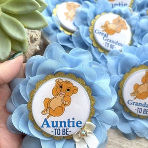 Pu&ograve; includere: Spilla a forma di fiore in tessuto azzurro con bordo glitterato dorato e grafica di Simba. La spilla riporta la scritta "Auntie -TO BE-" in blu. Un fiocco bianco con un cristallo &egrave; attaccato al fiore.