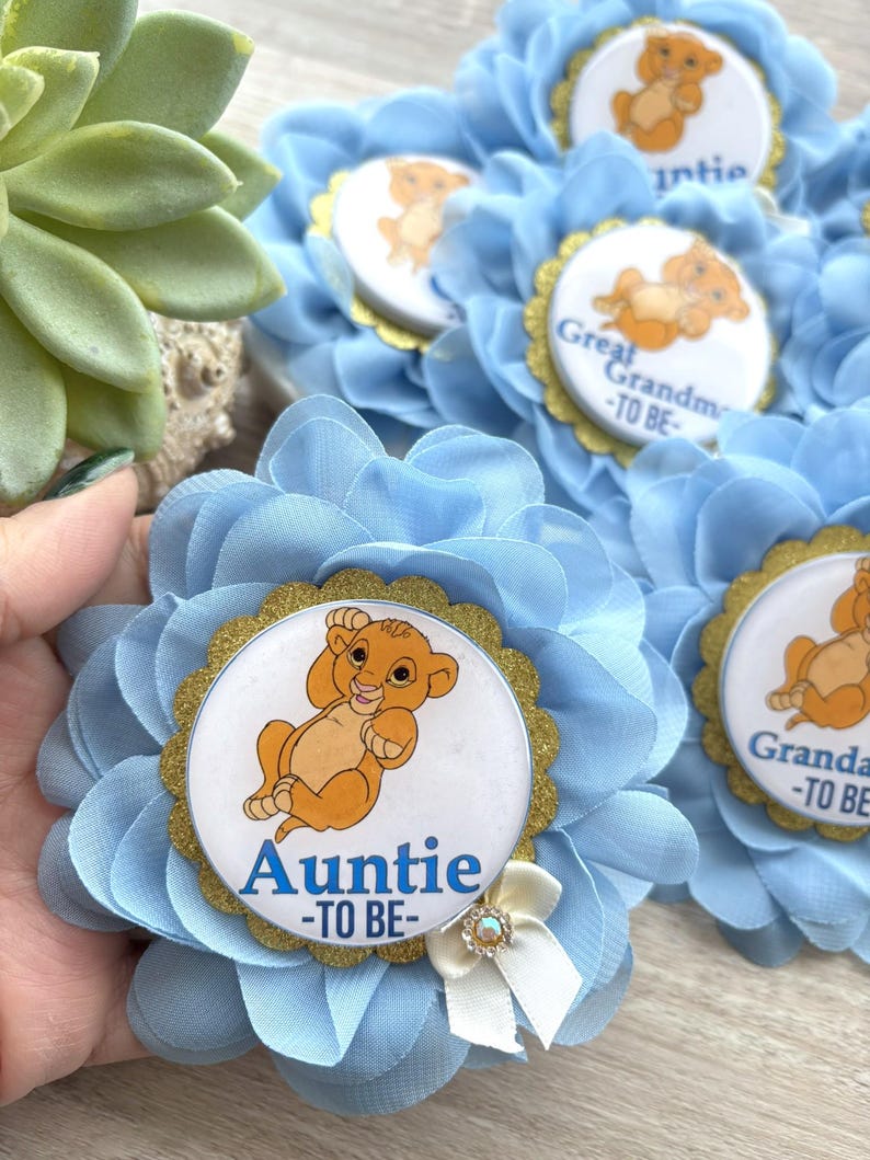 Pu&ograve; includere: Spilla a forma di fiore in tessuto azzurro con bordo glitterato dorato e grafica di Simba. La spilla riporta la scritta "Auntie -TO BE-" in blu. Un fiocco bianco con un cristallo &egrave; attaccato al fiore.