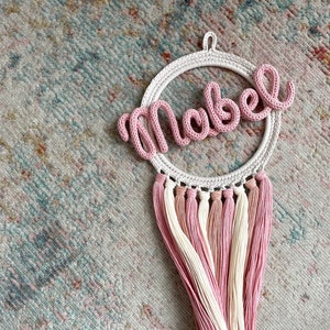 May include: A pink crocheted name sign with the name "Mabel" hanging from a white crocheted hoop with pink and white tassels.