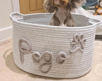 May include: A white and gray rope dog bed with the name "Roxy" embroidered on the side. A brown and white dog is sitting inside the bed.