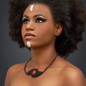 Puede incluir: Una mujer con un peinado afro y pintura facial blanca lleva un collar negro con un colgante de piedra roja.