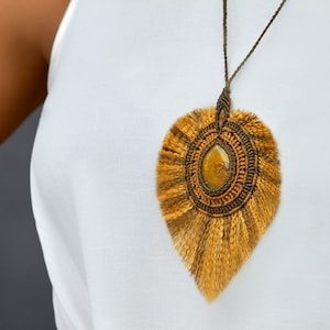 May include: A brown and gold macrame necklace with a large, teardrop-shaped pendant featuring a yellow gemstone. The pendant is surrounded by a fringe of brown and gold threads.