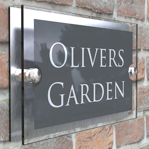 Puede incluir: Un letrero de n&uacute;mero de casa de vidrio transparente y plateado con el texto "OLIVERS GARDEN" en letras blancas sobre un fondo gris.