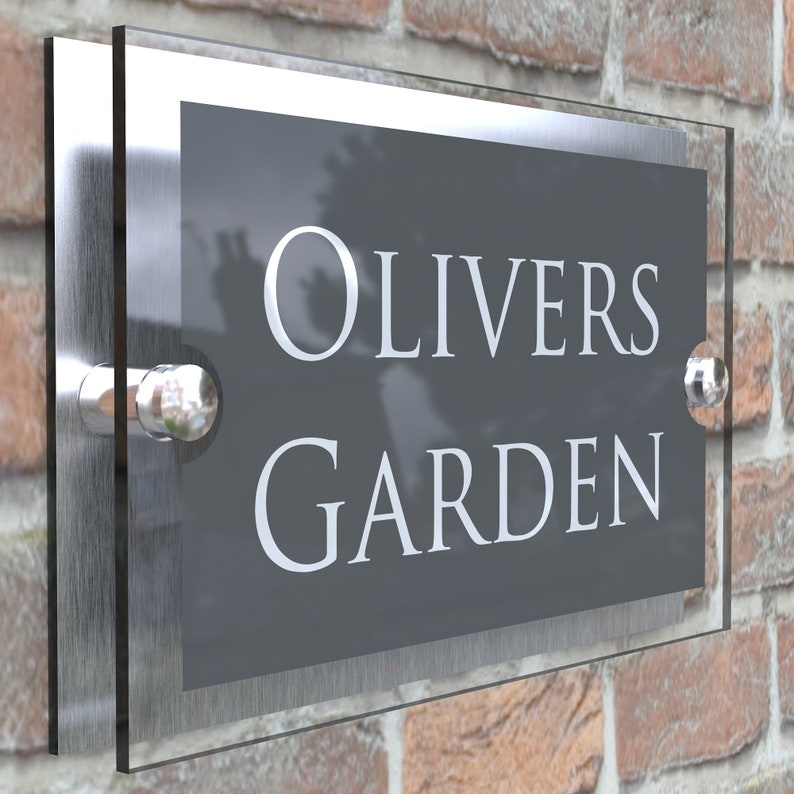 Puede incluir: Un letrero de n&uacute;mero de casa de vidrio transparente y plateado con el texto "OLIVERS GARDEN" en letras blancas sobre un fondo gris.