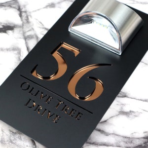 May include: Black house number sign with a copper-colored 56 and the text "Olive Tree Drive" in white. A small, silver, solar-powered light is mounted above the number.