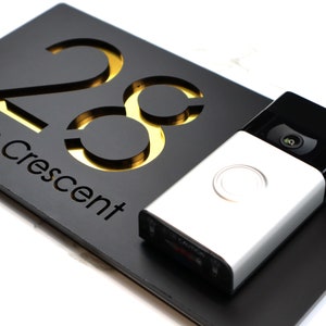 May include: A black and gold house number sign with the number 28 and the word "Crescent" below it. A silver doorbell camera is mounted on the sign.