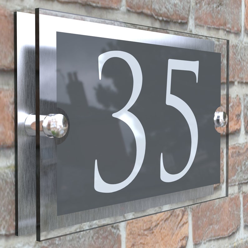 Puede incluir: Una placa de n&uacute;mero de casa de vidrio gris con el n&uacute;mero 35 en blanco. La placa est&aacute; montada en una pared de ladrillo.