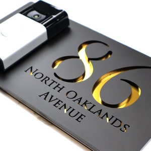 May include: A black house number sign with gold lettering. The number 86 is cut out of the sign and the gold lettering is behind the cut out. The sign also says "North Oaklands Avenue".